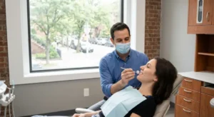 A professional dental examination in a modern Brooklyn clinic. A dentist wearing a surgical mask and a blue button-down shirt uses a small dental mirror to inspect a female patient's teeth. The room features a clean, neutral aesthetic with an exposed brick wall and a large window overlooking a city street, emphasizing a calm and trustworthy environment.